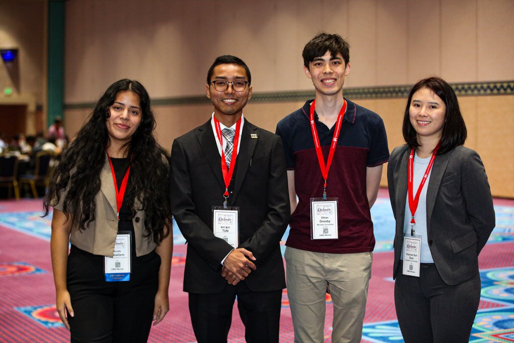 Four smiling students with conference badges posing together in a convention hall.