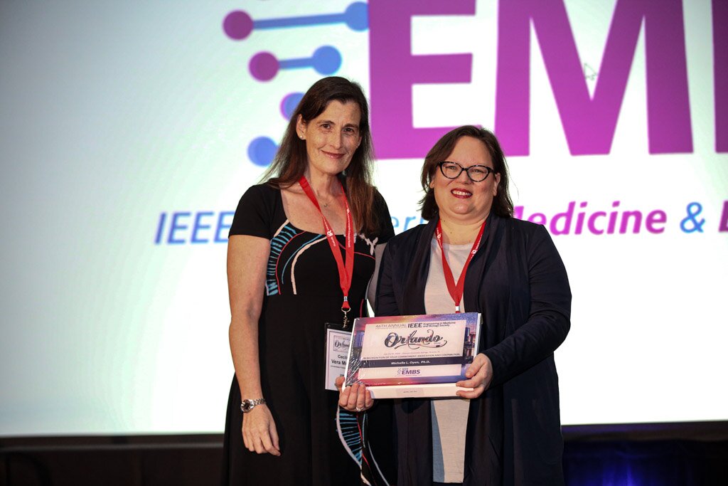 Two women posing with an award plaque in front of an IEEE EMBS screen.