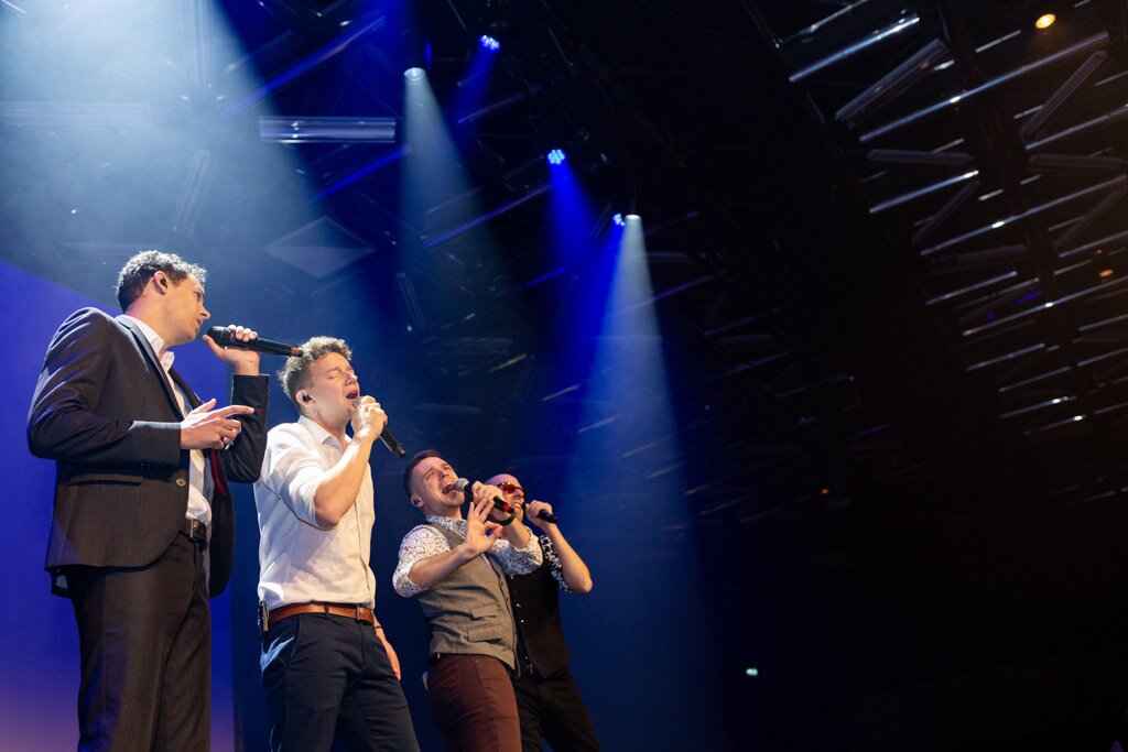 Four singers performing on stage under blue spotlights.