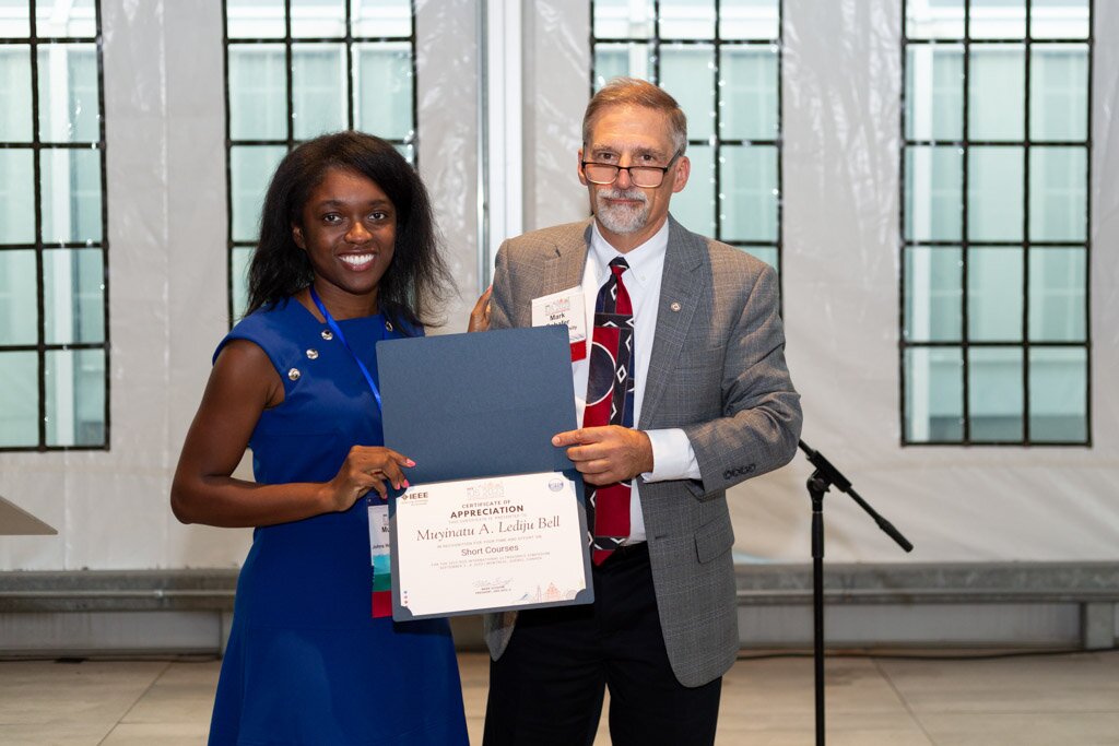 A woman smiling as she receives a certificate of appreciation from a man at a conference.