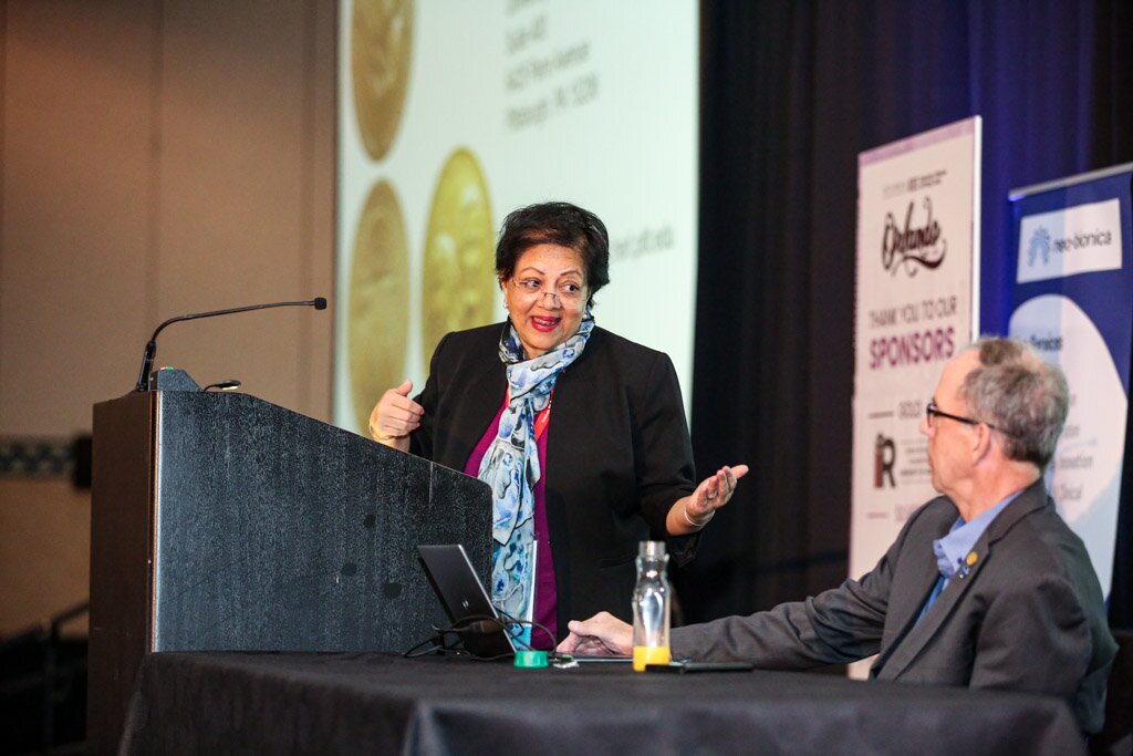 A woman speaking at a podium beside a seated colleague during a conference session.